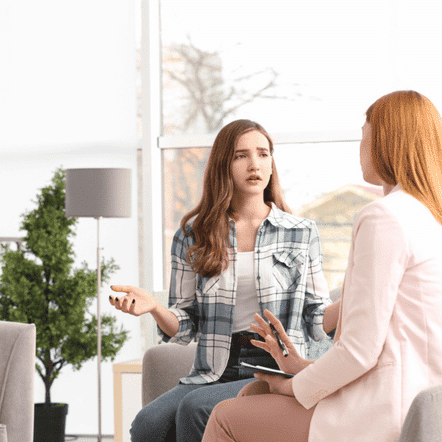 In a bright room, two women chat animatedly. The woman on the left gestures expressively, as if discussing something worth having, while the other listens intently. A large window and potted plants add tranquility to this scene that could grace any coaching website.