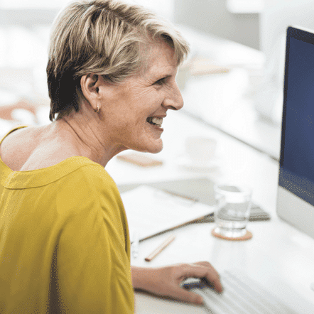 A woman with short blond hair and a yellow sweater is smiling at her computer screen, immersed in online marketing strategies for coaches. Around her, the blurred workspace reveals various office items that inspire productivity and creativity.