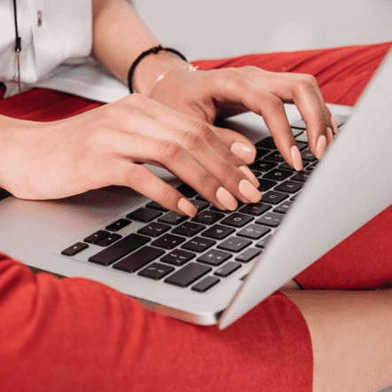 A person wearing red pants is typing on a silver laptop placed on their lap, hands poised over the keyboard, as they update content for their successful coaching website.