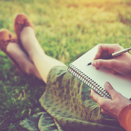 A person sits on the grass, penning thoughts in a spiral notebook, perhaps pondering how to write their first blog post. With a green patterned dress and brown shoes, they are gently illuminated by the soft sunlight.