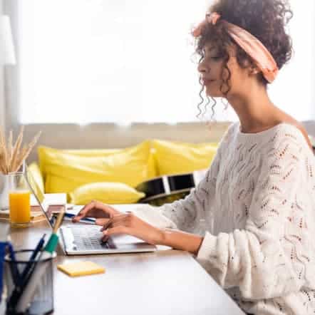 A woman with curly hair and a headband sits at a desk learning how to write her first blog post using a laptop. She wears a white sweater, with a notebook, pen holder, and glass of orange juice nearby. A yellow couch glows in the window's light behind her.