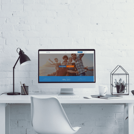 A modern workspace features a white desk and chair, a desktop computer showcasing a website redesign with a group photo, desk lamp, stationery, potted plant, and coffee mug. The setup is sleekly arranged against a white brick wall backdrop, creating an inspiring place for productivity.