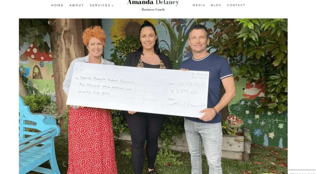 Amanda and Ian Delaney stand  in front of a mural. They are holding   a large check for €2,375, dated 24/05/2023, made out to Saoirse Domestic Violence Services. They are casually dressed and smiling.