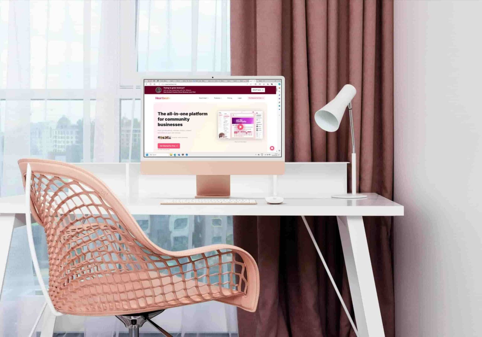 A modern workspace featuring a white desk with a computer displaying the heartbeat.chat community platform. There's a beige, intricately designed chair, a small lamp, and a window with brown curtains in the background. Soft natural light fills the room.