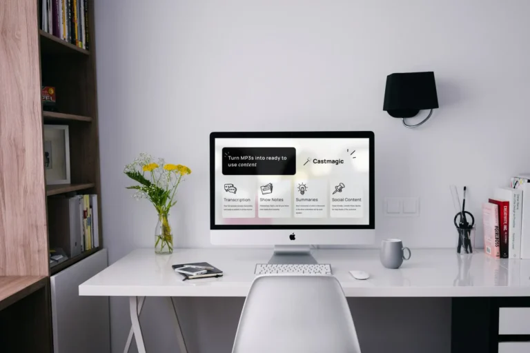 A modern desk setup showcasing an Apple computer with Castmagic software for transcription, show notes, summaries, and social content. A vase with yellow flowers, a lamp, and books are neatly arranged on the white desk.