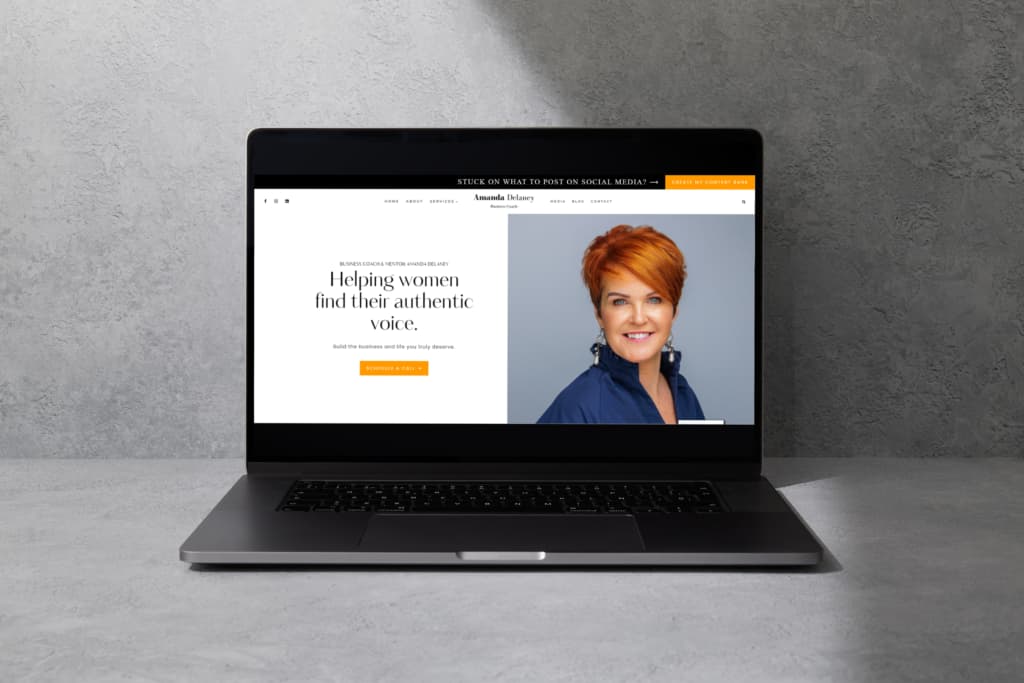 A laptop displays a website featuring Business Coach Amanda Delaney - a smiling woman with short red hair. The headline reads, "Helping women find their authentic voice." The background is a textured grey wall, highlighting the site's responsive web design for optimal viewing on any device.