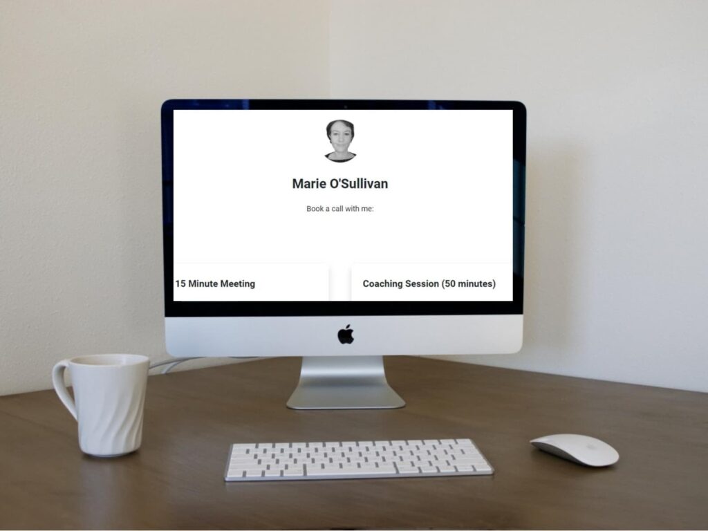 A computer screen shows a scheduling page for booking calls with someone named Marie O'Sullivan, offering options for a 15-minute meeting or a 50-minute coaching session. A white mug and a computer mouse are on the wooden desk.