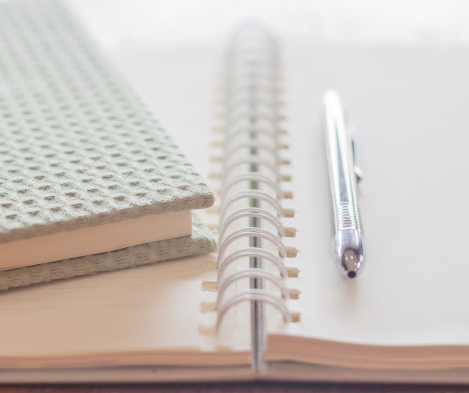 An open spiral notebook lies on a table with a silver pen placed on top. Beside it is a closed textured notebook with a light green cover. The scene is softly lit, conveying a calm and organized workspace.