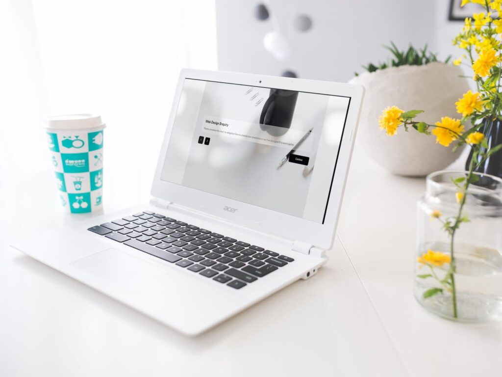 A white Acer laptop on a table showcases a webpage, highlighting the web design process. Beside it, there's a cup with a blue pattern and a glass jar with yellow flowers. In the background, a soft-focused white wall and a plant in a white pot complete the scene.