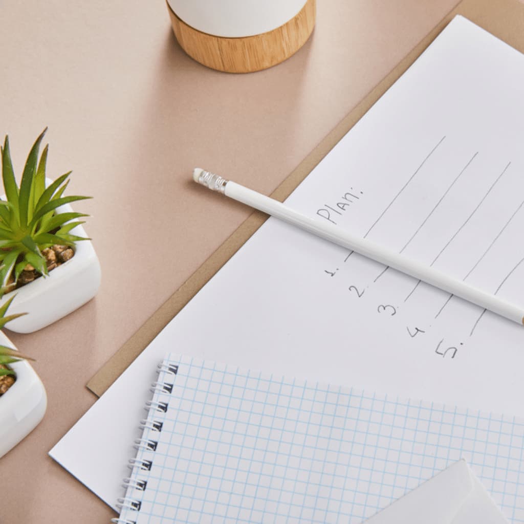 A top view of a desk with a notepad and a sheet labelled "Plan" featuring a list numbered 1 to 5 for an upcoming website planning workshop. A pencil lies ready on the paper. Two small potted plants add greenery on the side, and part of a steaming cup peeks from the corner.