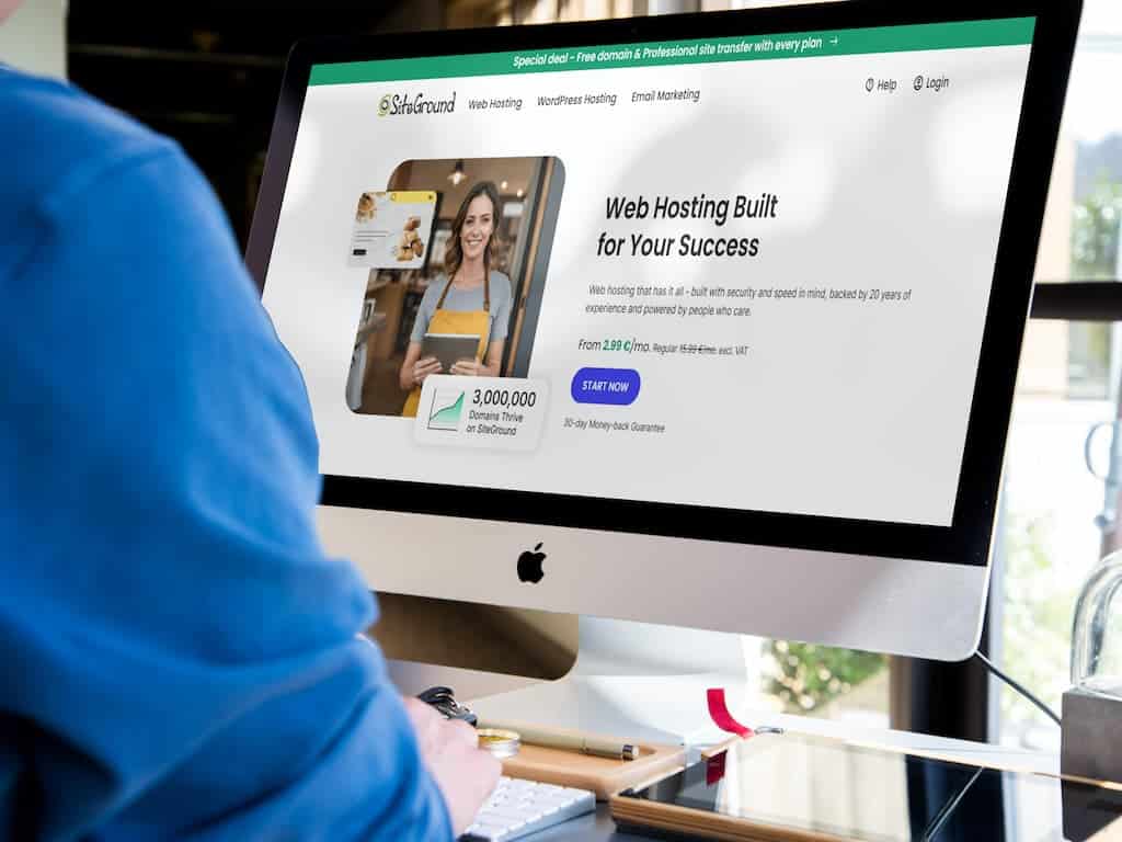 A person wearing a blue sweater is sitting at a desk, navigating the Siteground web hosting service website on a large desktop computer. The screen displays promotional content and pricing details about web hosting plans.