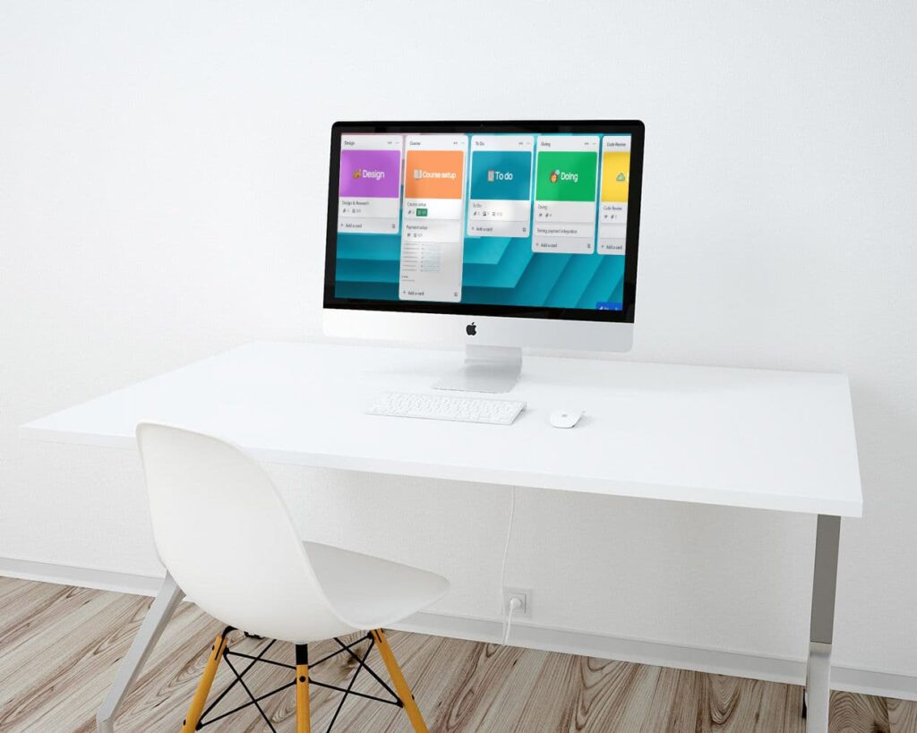 A minimalist workspace highlights the web design process with an iMac displaying colourful task cards on a sleek white desk. A white keyboard and mouse sit neatly beneath, while a modern white chair invites creativity. The wooden floor and plain white wall complete the clean, focused environment.