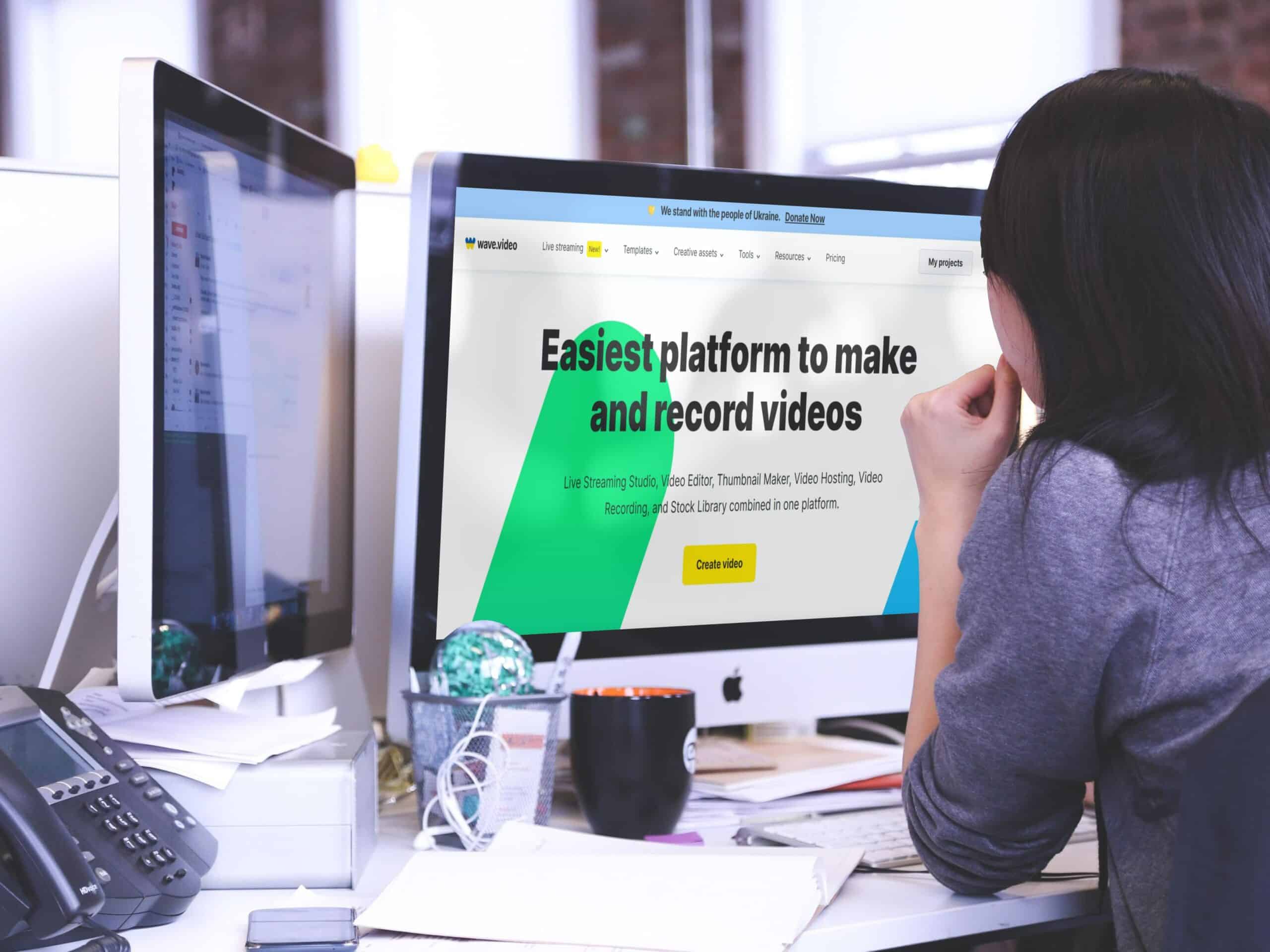 A person sitting at a desk, looking at a computer screen displaying the Wave.video website with the text "Easiest platform to make and record videos." The desk has a keyboard, a smartphone, and a coffee mug.