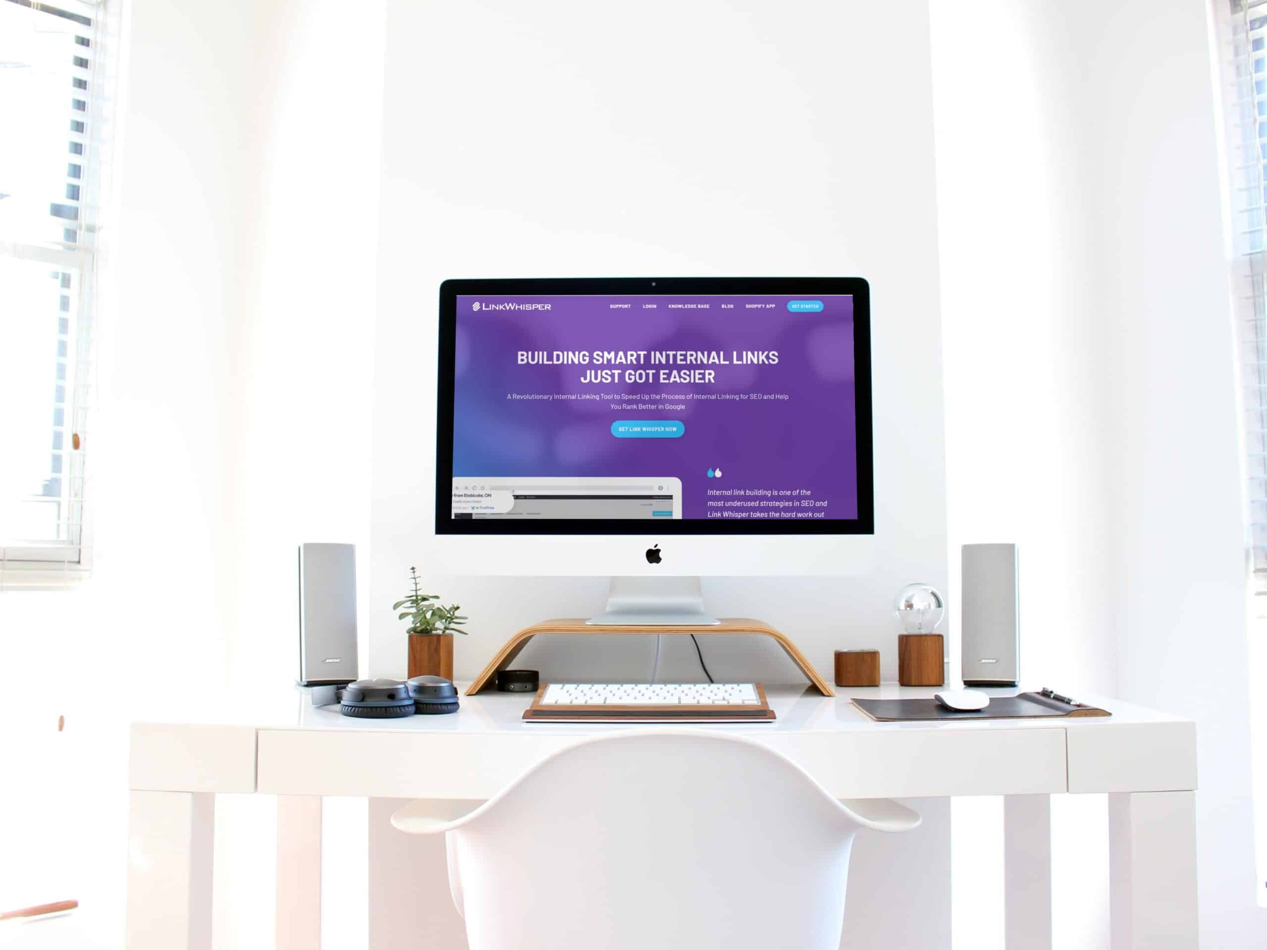 A minimalist workspace with an iMac displaying the Link Whisper website that reads "Building Smart Internal Links Just Got Easier." The desk includes speakers, a plant, a mouse, and a keyboard, all set in a bright room with large windows.